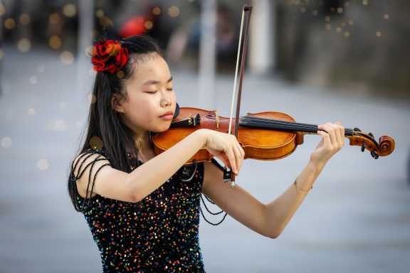 Ein Mädchen spielt Violine, trägt ein glitzerndes Kleid und hat eine Blume im Haar.